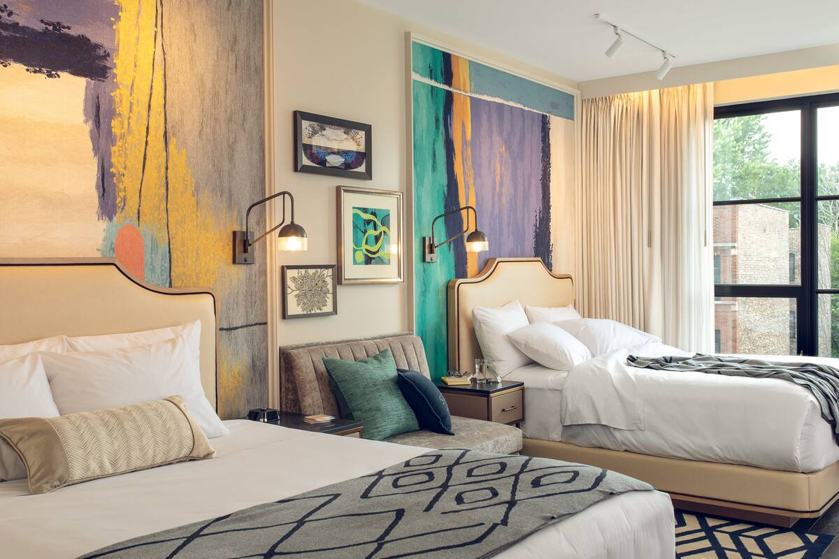Quarto de hotel com duas camas, uma janela grande e obras de arte nas paredes.