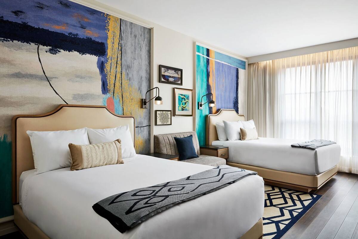 Quarto de hotel com duas camas, um quadro abstrato grande, uma mesa de cabeceira e uma janela com cortinas.