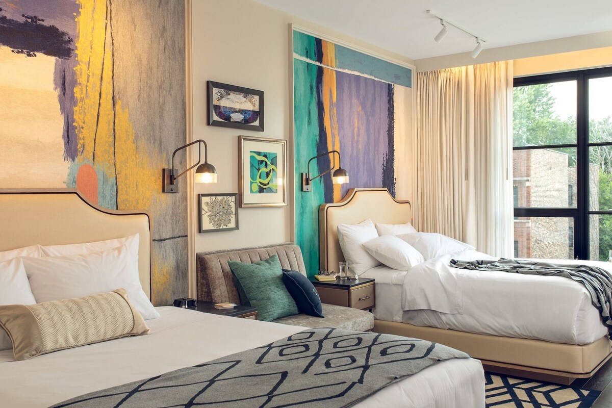 A hotel room with two beds, a large window, and artwork on the walls.