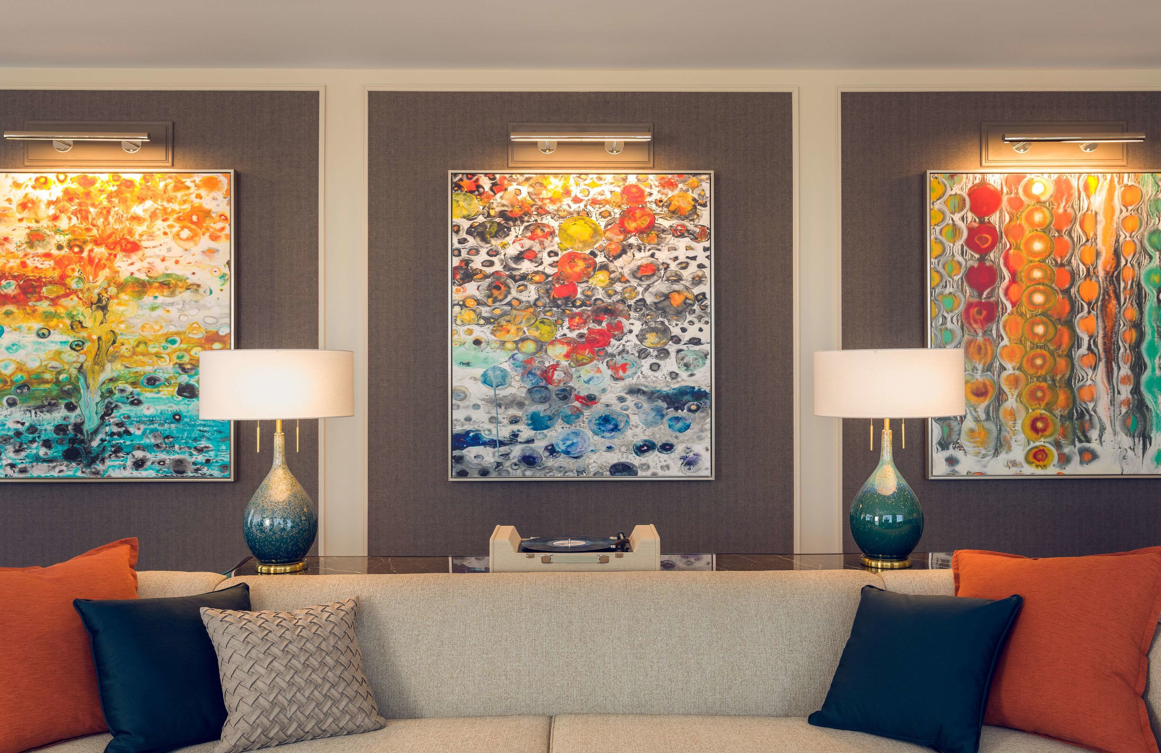 A modern living room with a beige sofa, two side tables with lamps, and three colorful abstract paintings on the wall.