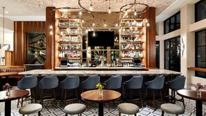 Una zona de bar con mostrador de mármol, varias estanterías llenas de botellas y una gran pantalla de televisión. Hay varios taburetes de bar y una mesa redonda con un jarrón de flores.