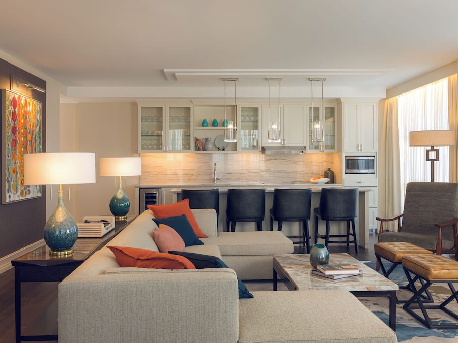 A modern living room with a sofa, ottoman, and coffee table, a kitchen area with bar stools, and a dining table.