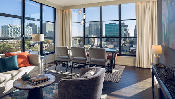Una habitación de hotel moderna con ventanas amplias, un sofá, sillas, una mesa de comedor y una mesa de centro.