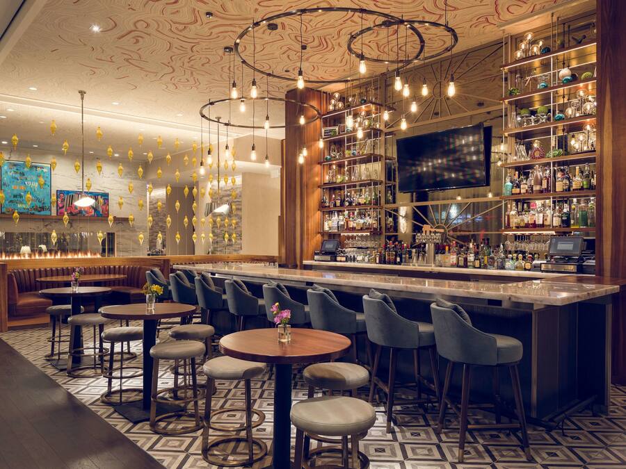 A bar area with multiple seating options, including round tables and bar stools. There are shelves with bottles and glasses, and a television screen is mounted on the wall.