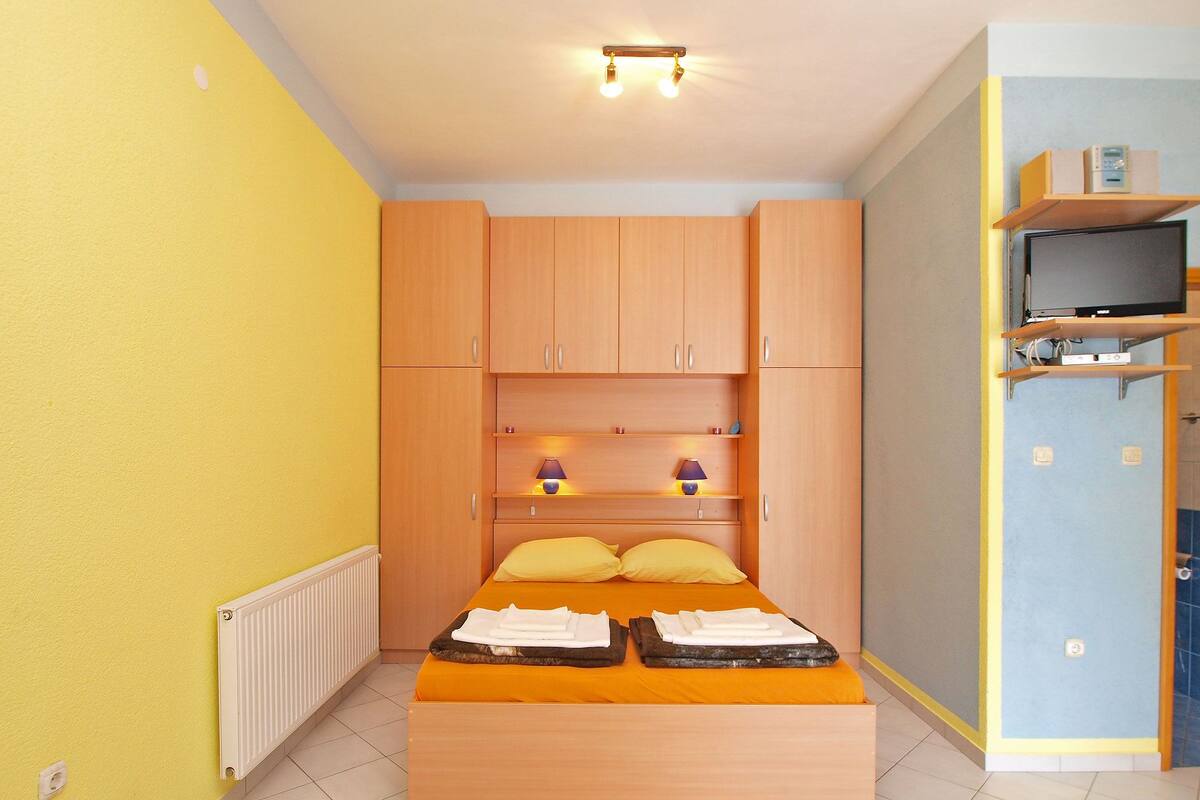 A compact bedroom with a bed, wooden cabinets, a shelf with a television, and a radiator.
