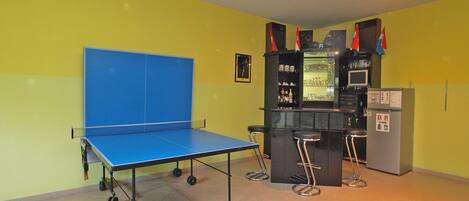 A room with a ping pong table, a bar area with stools, and a refrigerator.