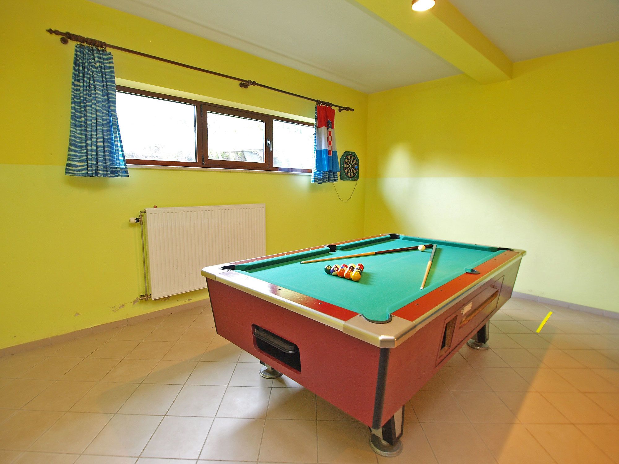 Una mesa de billar en una habitación con paredes amarillas, una ventana con cortinas azules y un dardo en la pared.