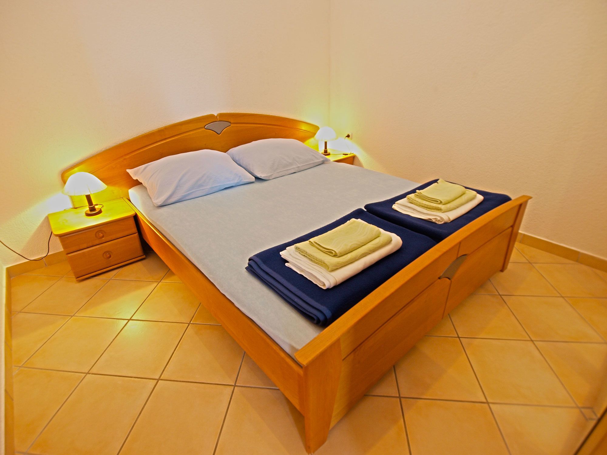 Een tweepersoonsbed met houten frame, twee kussens, een nachtkastje met lamp en twee opgevouwen handdoeken op het bed.