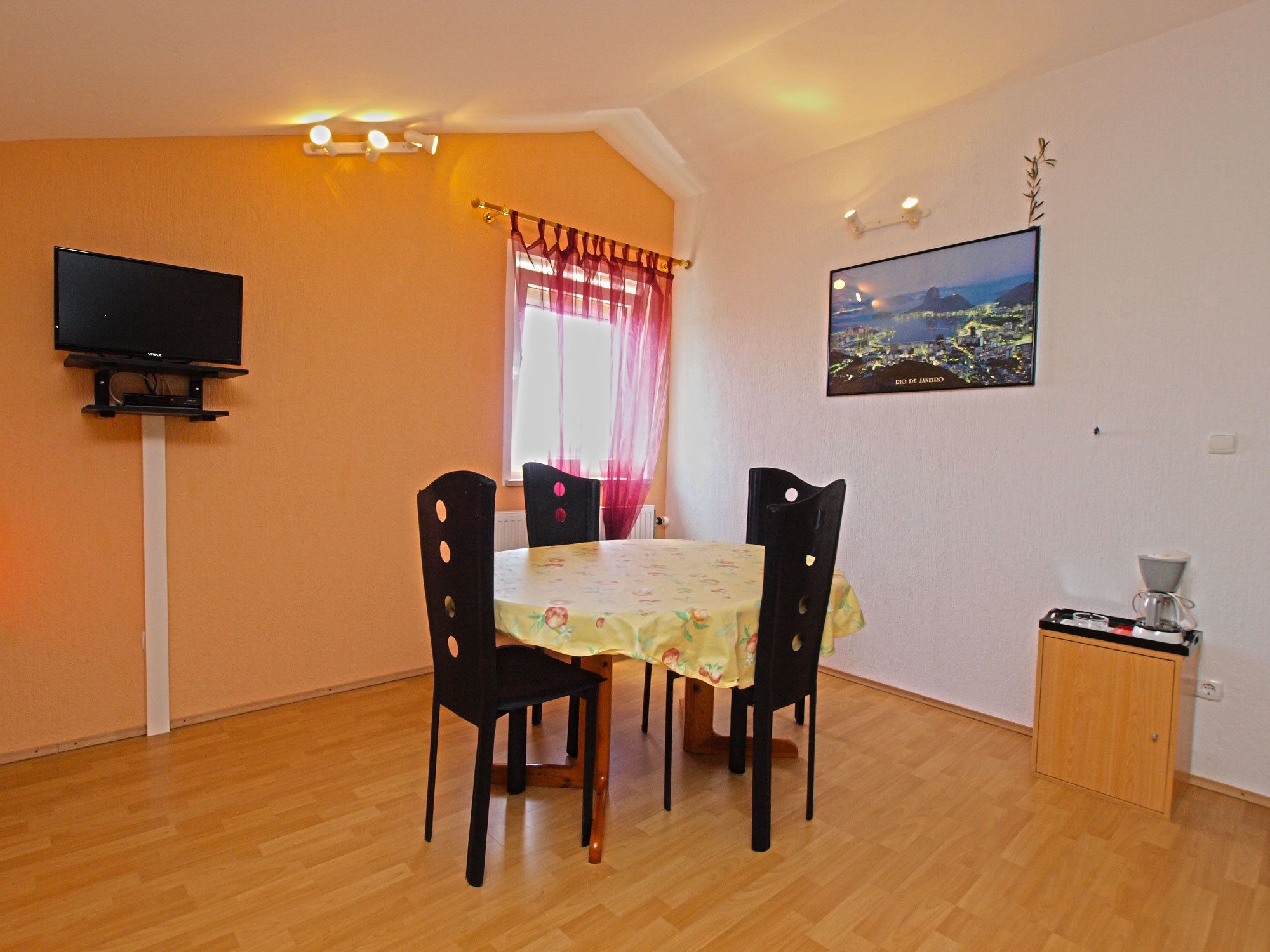 A dining area with a table and four chairs, a television on a stand, a window with curtains, a picture on the wall, and a small cabinet with a coffee maker.