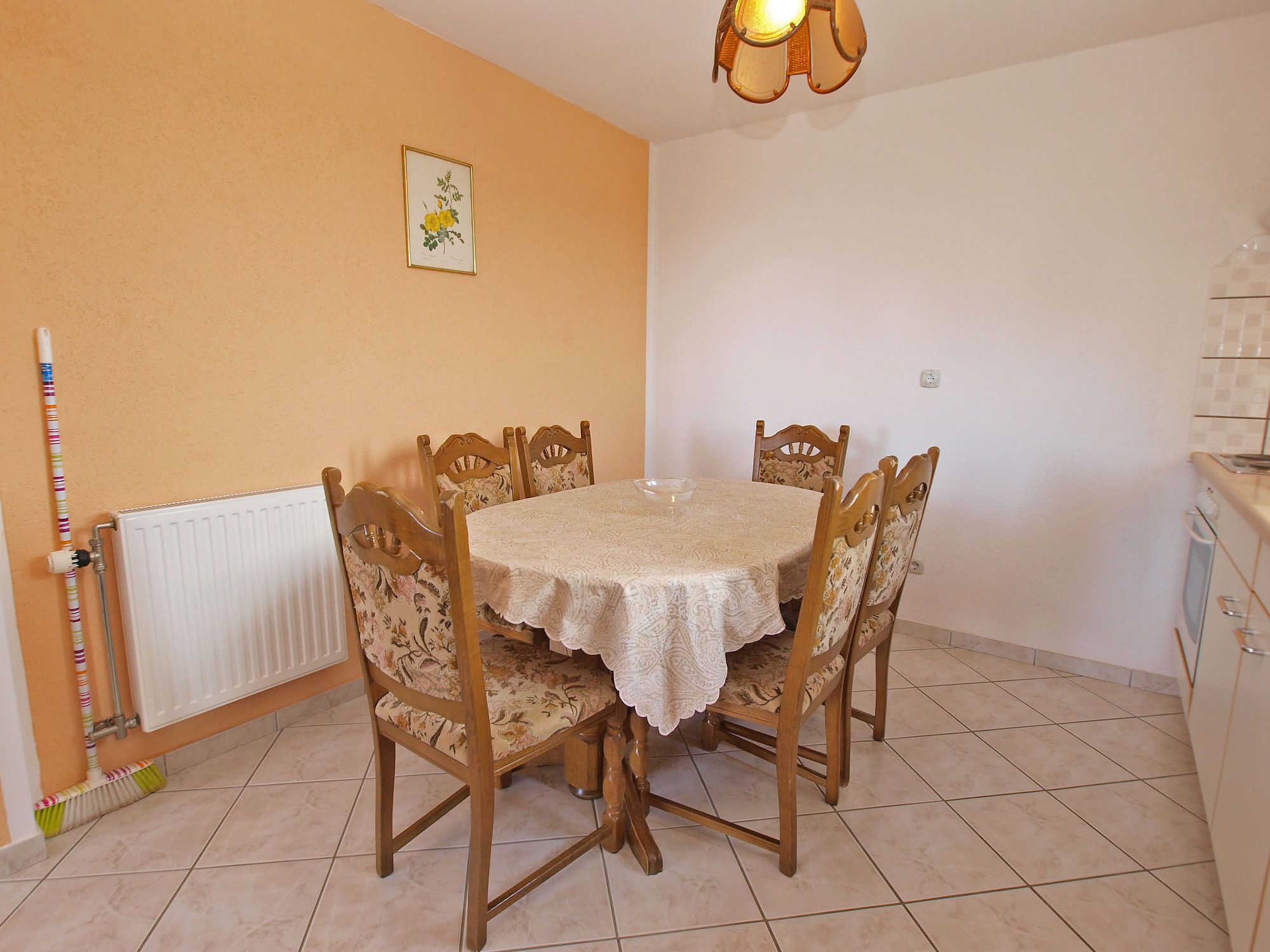 Een eetgedeelte met een ronde tafel bedekt met een kantkleed, omringd door houten stoelen met bloemenmotieven. Een radiator is links zichtbaar, en een wandlamp hangt boven de tafel.