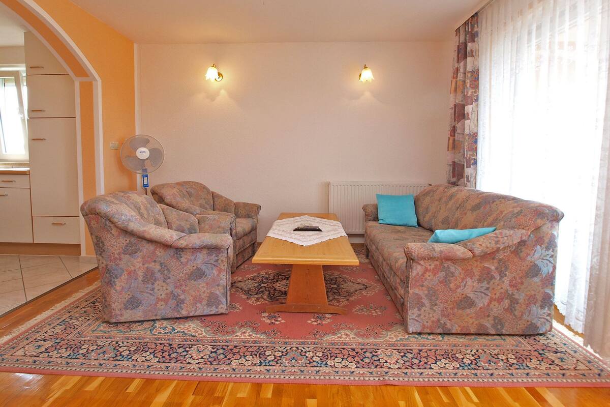 A living room with two patterned armchairs, a wooden coffee table, and a sofa with a blue pillow. There is a patterned rug on the floor, a wall-mounted fan, and two wall sconces.