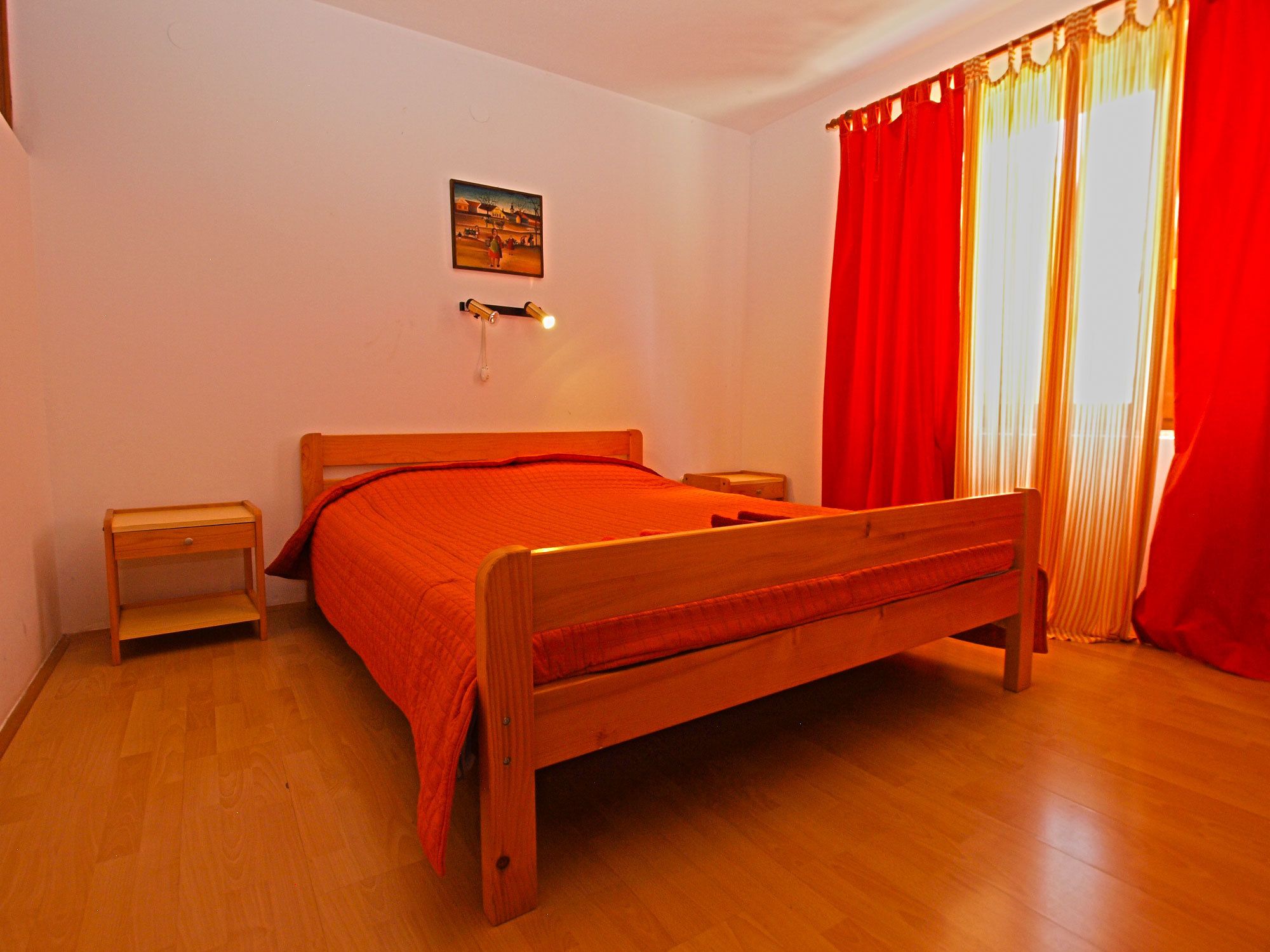 A bedroom with a wooden bed, a bedside table, a nightstand, a wall-mounted lamp, and a window with red curtains.