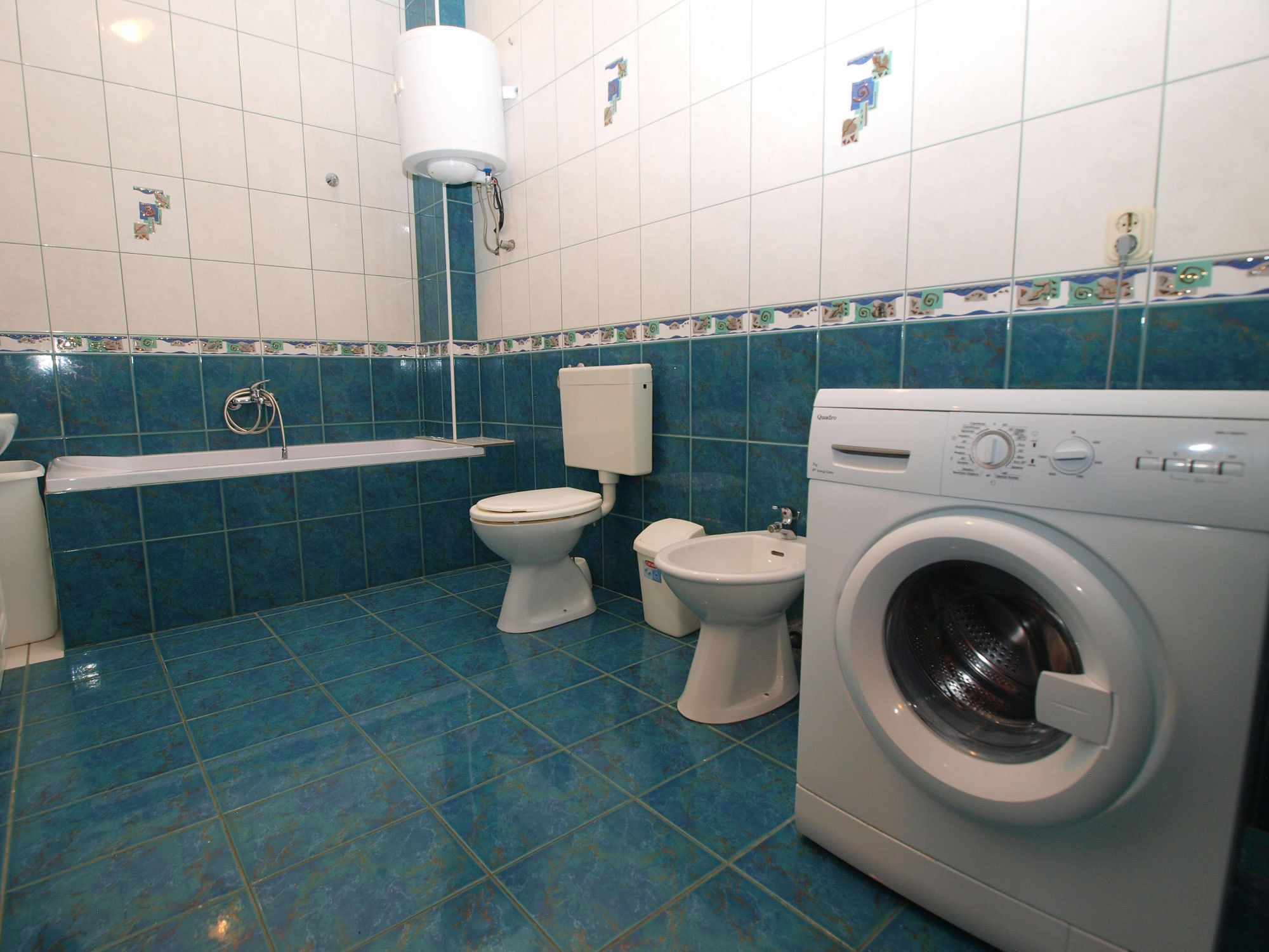 A bathroom with blue tiles, a toilet, a bidet, a washing machine, and a bathtub.