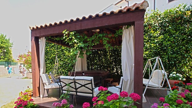 A covered outdoor seating area with a table and chairs, surrounded by lush greenery and vibrant flowers. - Apartment Marija 85 (Fazana)