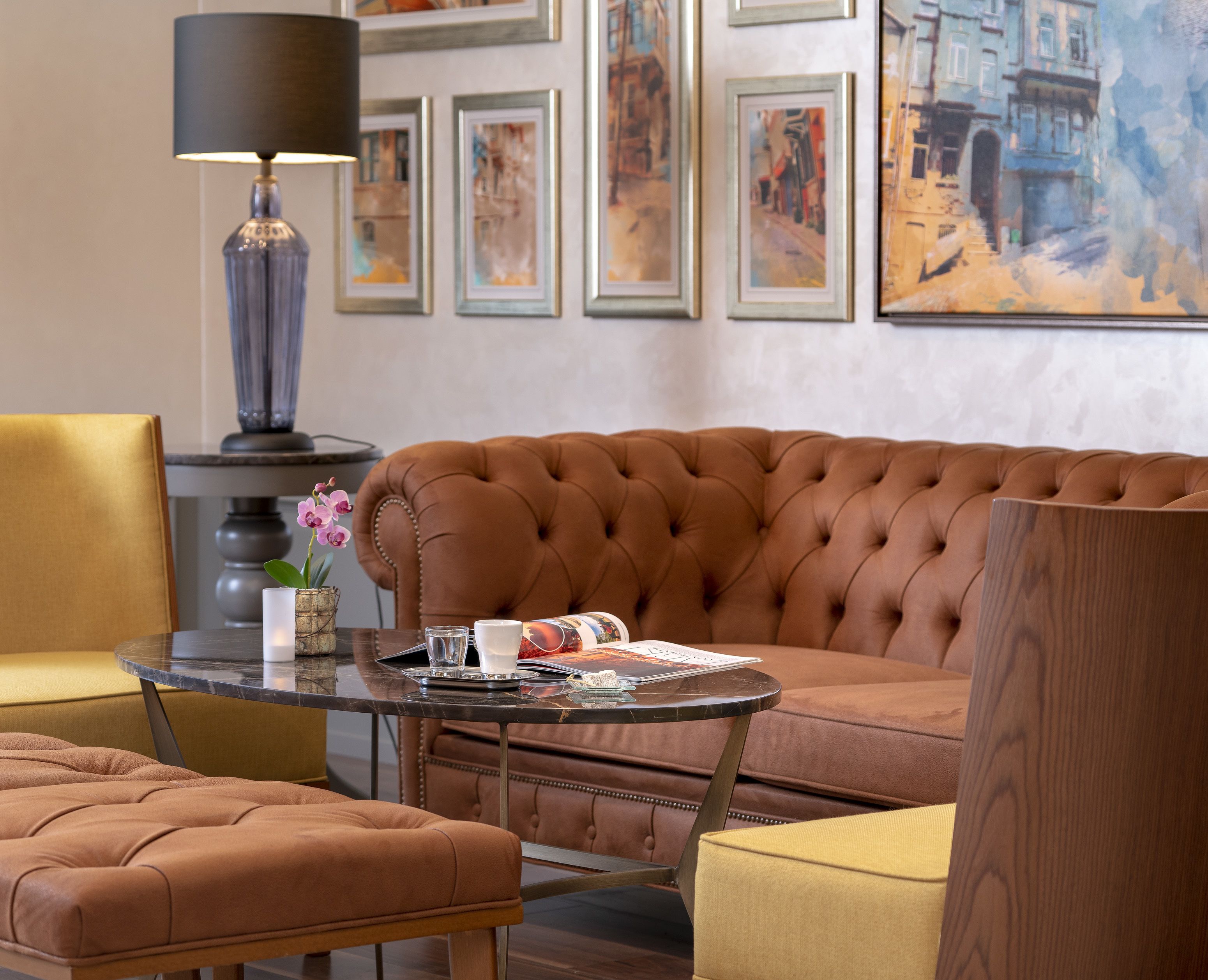 A modern living room with a tufted brown sofa, a round glass coffee table, and a side table with a lamp. There are framed artworks on the wall and a vase with flowers on the table.