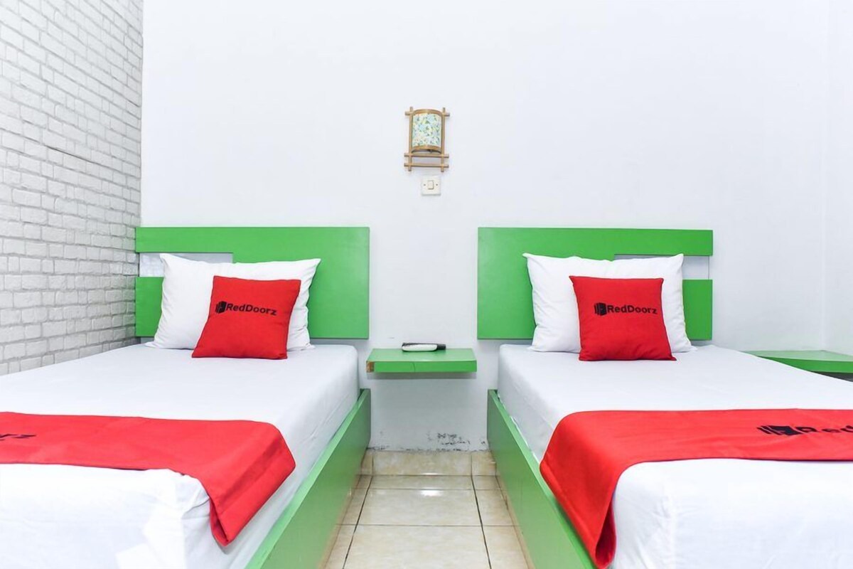 A hotel room with two beds, each with green headboards and white bedding. There is a small bedside table with a lamp between the beds.