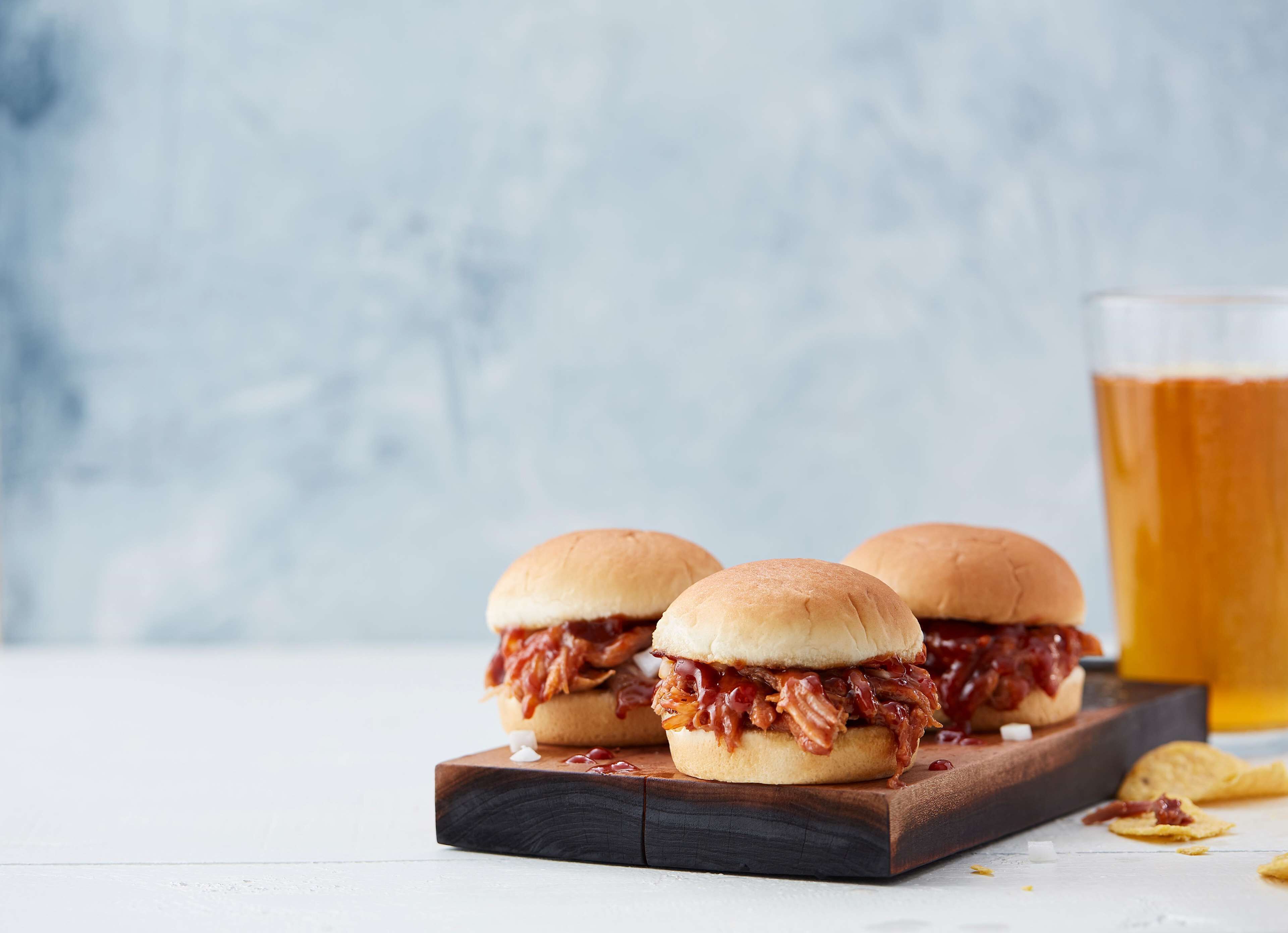 Dos sándwiches de cerdo desmenuzado sobre una tabla de madera, con una cerveza al fondo.