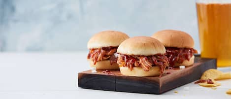 Two pulled pork sandwiches on a wooden board with a glass of beer in the background.