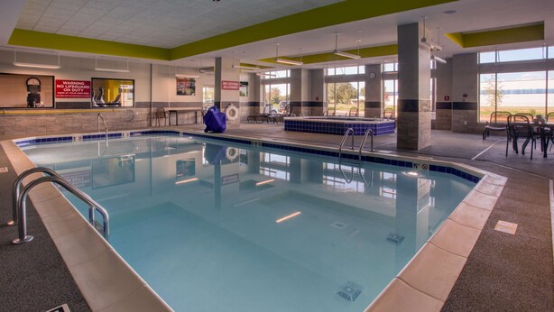 An indoor swimming pool with a 5 ft depth sign, a no diving warning, and a view of the outdoors through large windows.