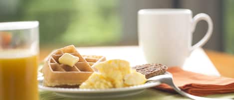 Desayuno con waffles, huevos revueltos, tostadas, un vaso de jugo de naranja y una taza blanca sobre una mesa con un mantel individual verde.