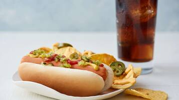 A hot dog with toppings and chips on a plate, with a drink in the background.
