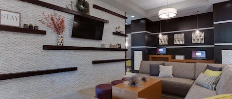 A modern hotel lobby with a brick-patterned wall, a 'STAY' sign, a flat-screen TV, and a seating area with sofas and a coffee table.