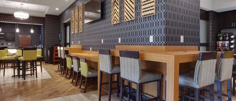 A modern bar area with wooden bar counters and striped chairs, a large TV on the wall, and a patterned wall design.