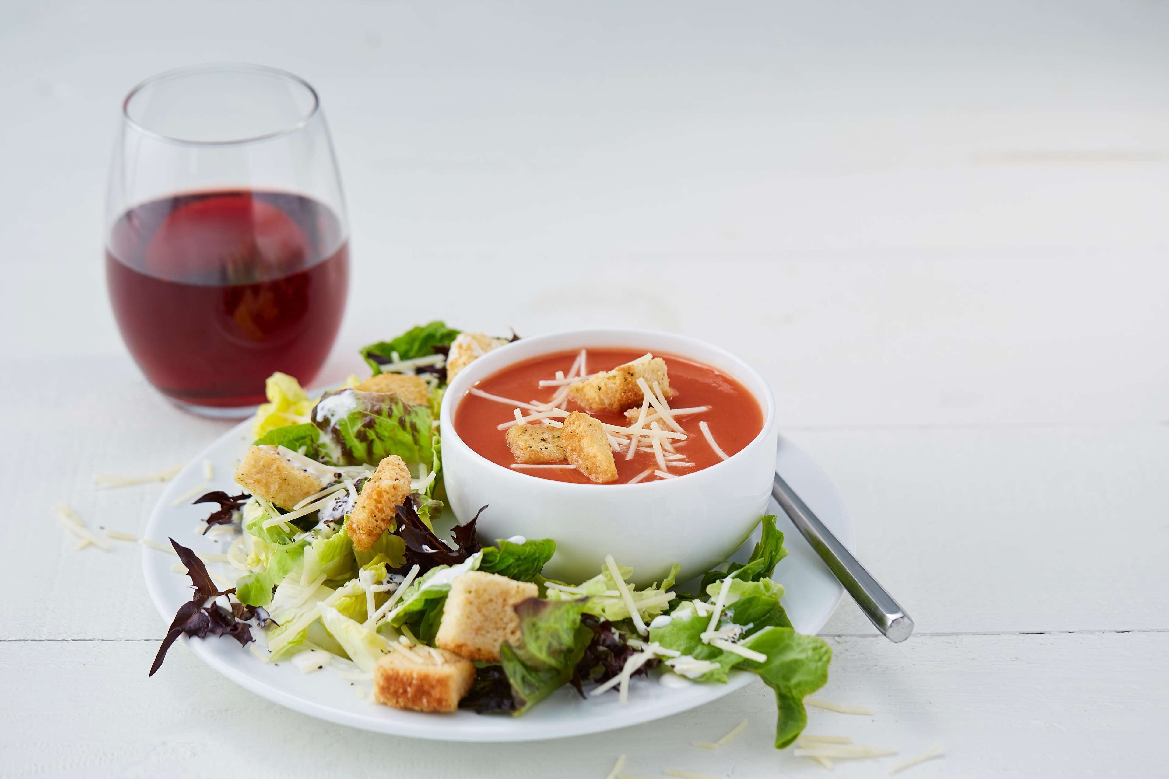 Un plato blanco con ensalada y un tazón de sopa de tomate, decorado con cubitos de pan y queso.