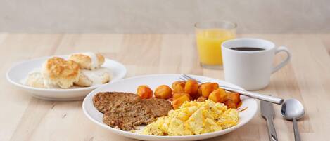 A breakfast plate with scrambled eggs, sausage, and roasted potatoes, a plate of biscuits, and a glass of orange juice.