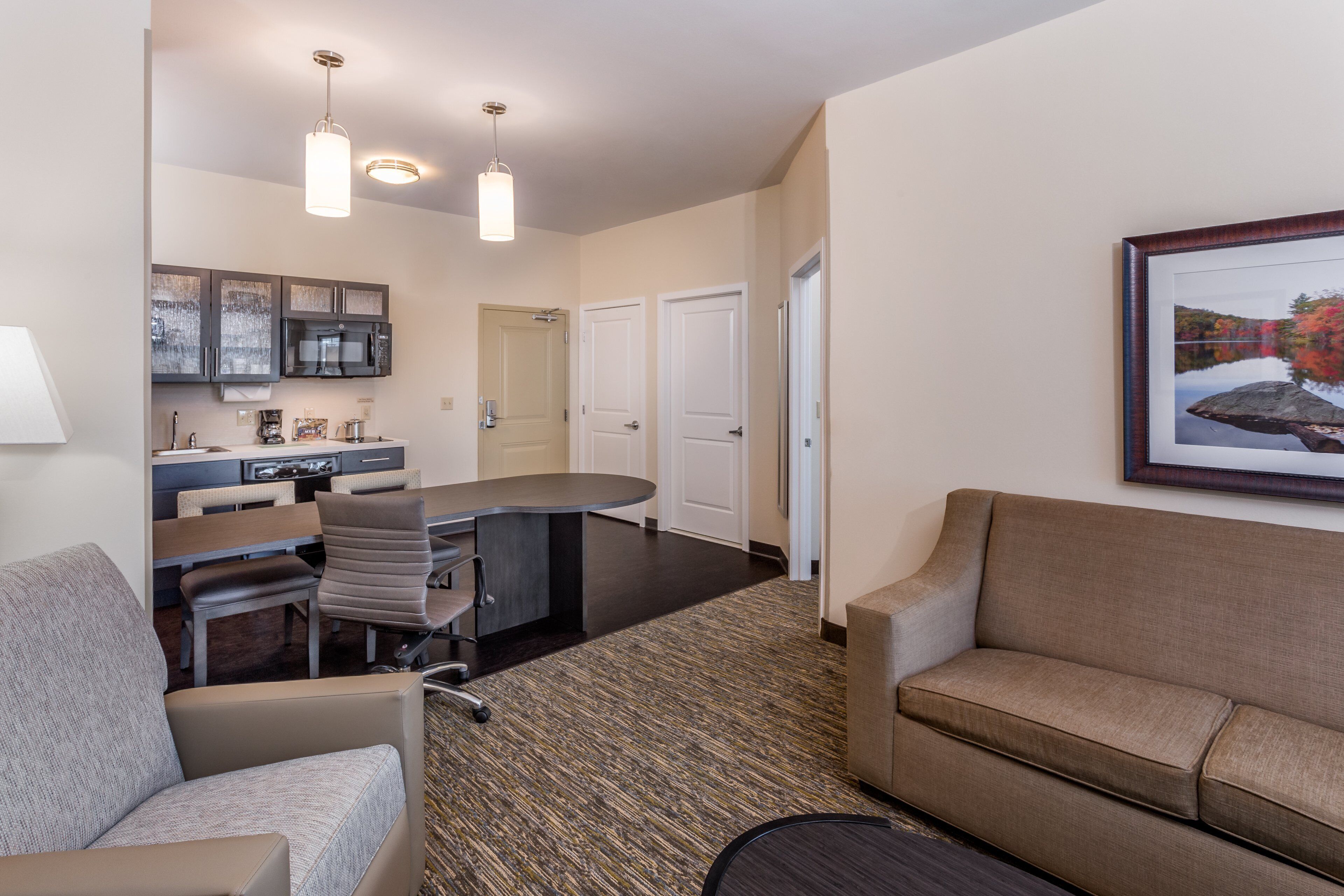 A modern hotel room with a kitchenette, a dining area, a sofa, and a framed picture on the wall.