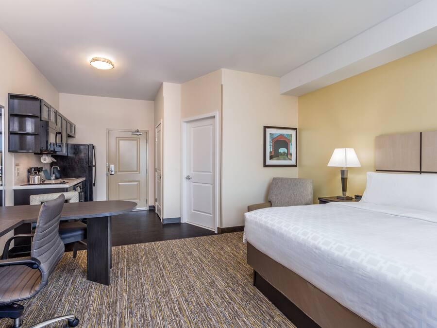 A hotel room with a bed, a desk with a chair, a kitchenette, and a door leading to another room.