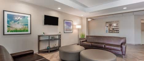 A modern living room with a brown leather sofa, two grey ottomans, a flat-screen TV, and framed artwork on the walls.