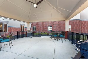 A covered patio area with outdoor seating, a ceiling fan, and a brick building in the background.