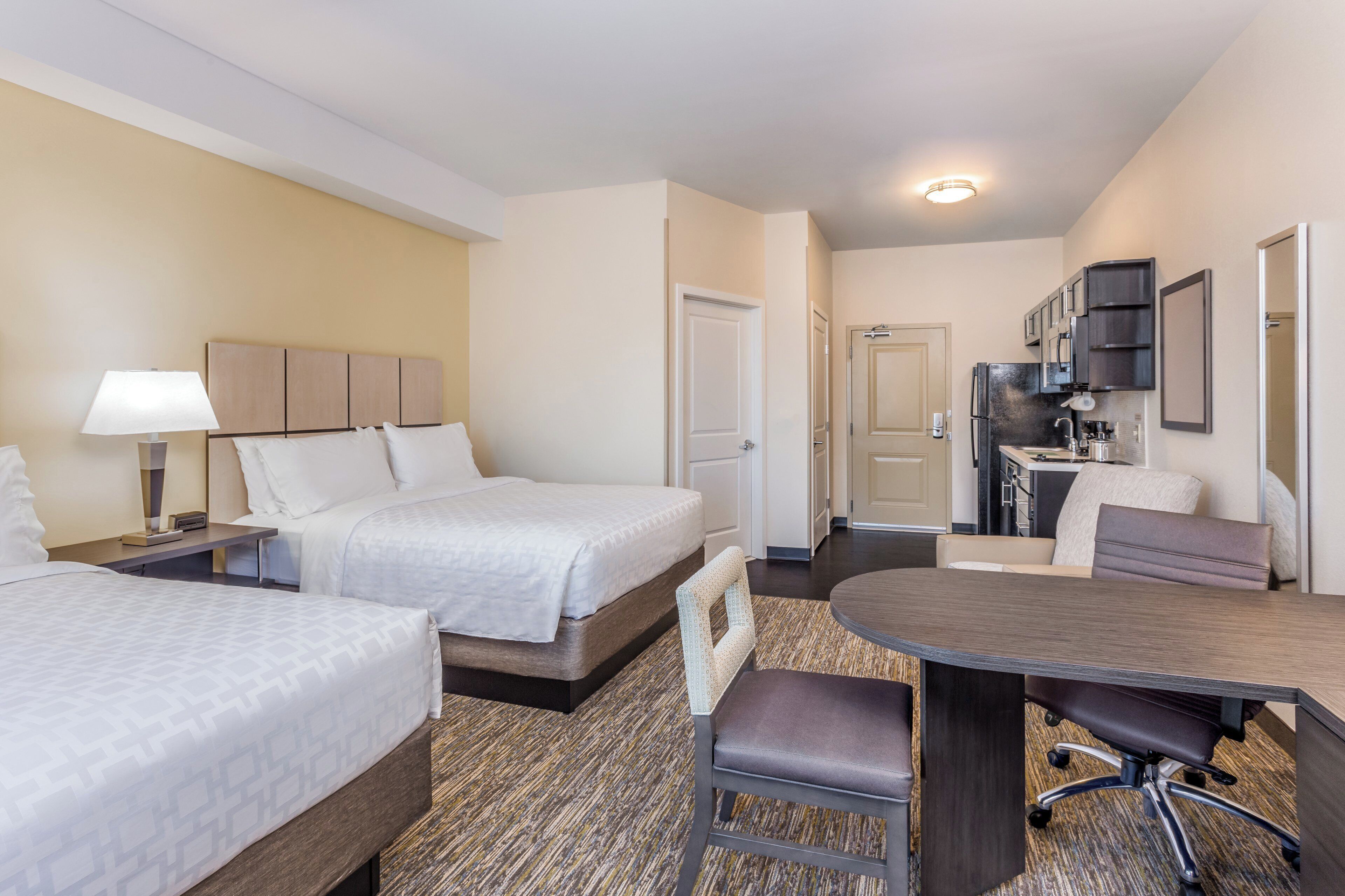A hotel room with two beds, a desk, a chair, a kitchenette, and a bathroom.