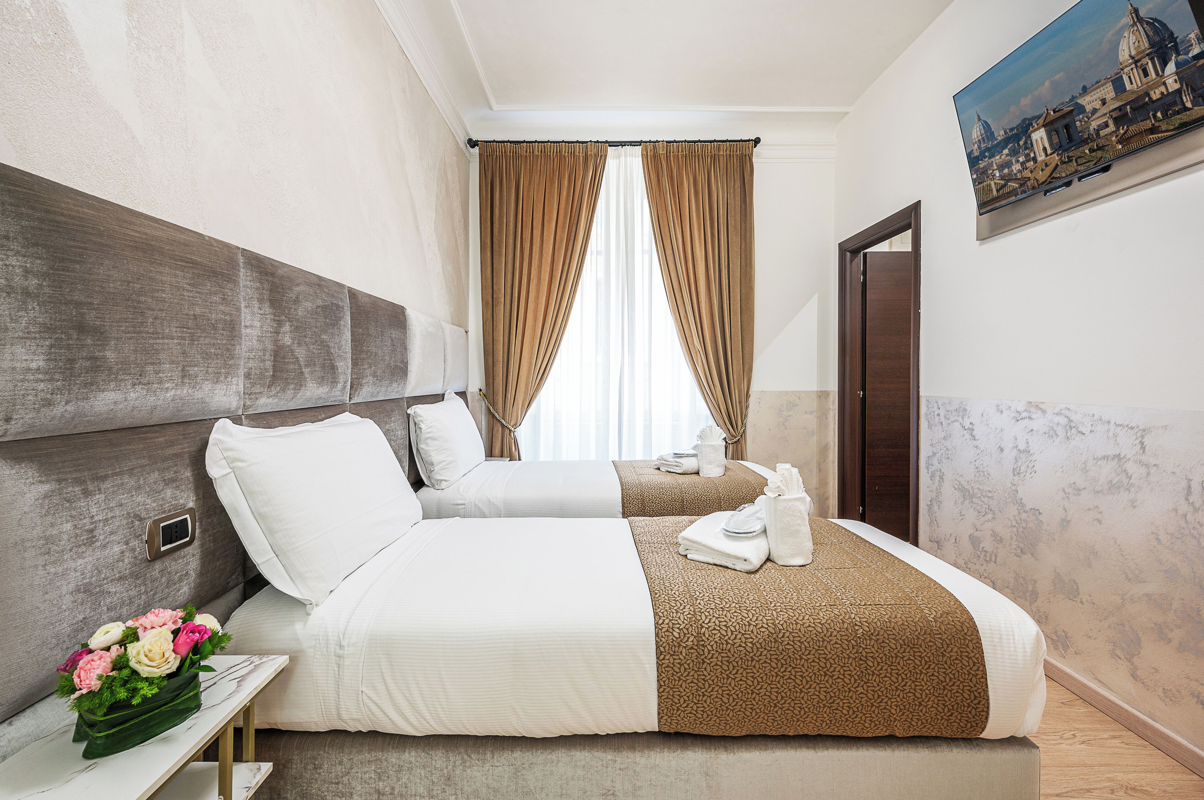 A hotel room with two beds, a small table with a vase of flowers, a painting on the wall, and a view of a cityscape.