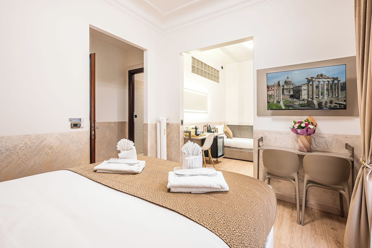 Habitación de hotel con cama, escritorio, silla, un cuadro y baño visible a través de una puerta abierta.