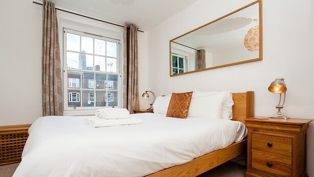 A neatly made bed with white linens, a wooden headboard, a nightstand with a lamp, and a large window with curtains. - Stylish One Bedroom Flat in Vauxhall (London)