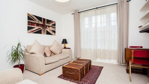 A living room with a beige sofa, a red chair, a table, a lamp, a potted plant, and a large window with curtains. - Stylish One Bedroom Flat in Vauxhall (London)