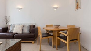 Uma sala de estar moderna com um sofá marrom, uma mesa de centro de vidro e uma mesa de jantar de madeira com quatro cadeiras. Há dois lustres fixados na parede e um quadro na parede.