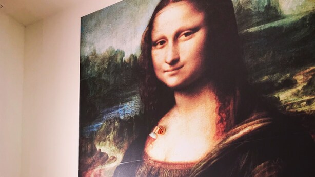 Un dormitorio con cama, almohadas, una mesita de noche, un florero con flores y un cuadro grande de la Mona Lisa en la pared.