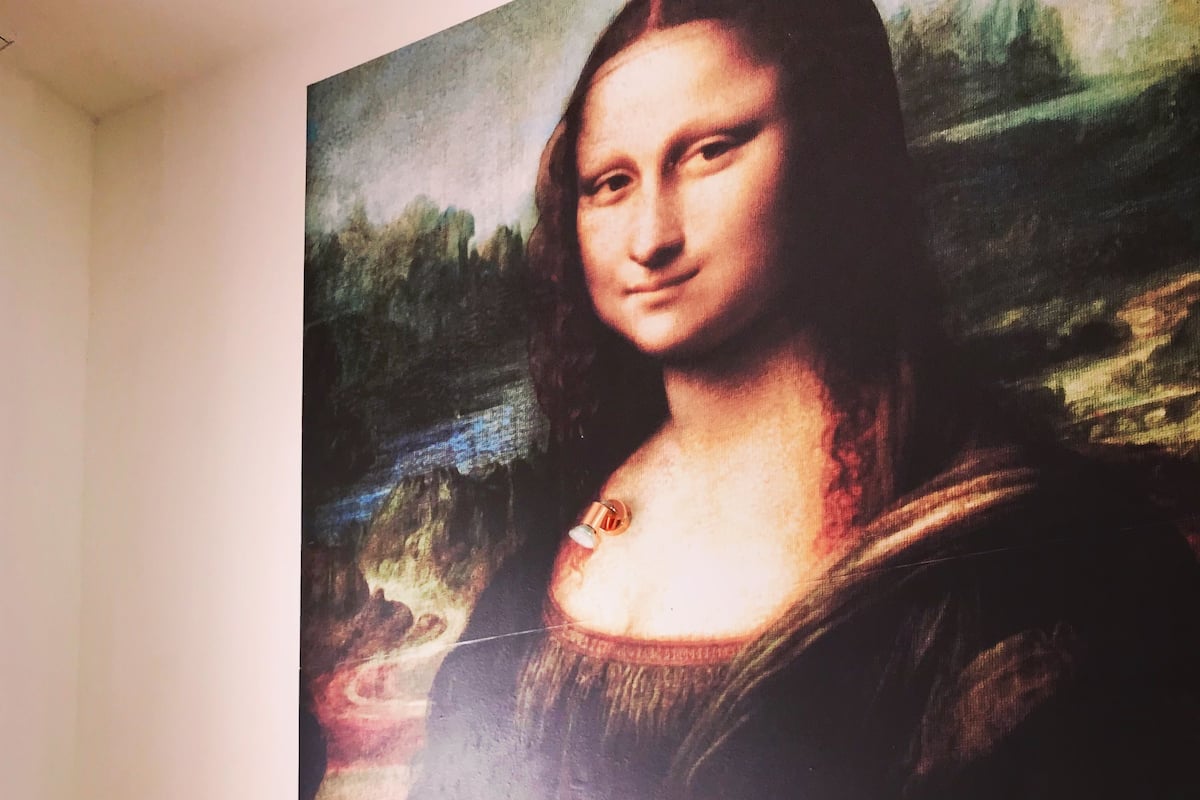 Ett sovrum med en säng, kuddar, ett nattduksbord, en vas med blommor och en stor målning av Mona Lisa på väggen.