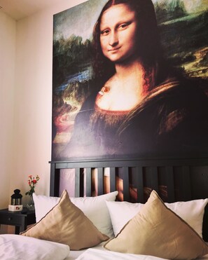 A bedroom with a bed, pillows, a nightstand, a vase with flowers, and a large painting of the Mona Lisa on the wall. - home2be apartments (Wuppertal)