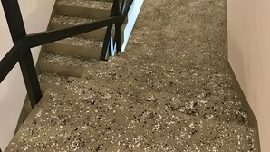 Una escalera con barandilla negra que lleva a una plataforma. El suelo está cubierto de baldosas con motas.