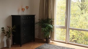 Una habitación con un aparador de madera oscura, una planta en maceta, un jarrón con flores y un ventanal con vistas a árboles.
