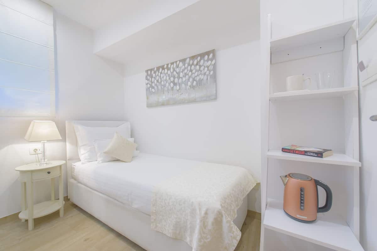 Quarto bem arrumado com cama, mesa de cabeceira, luminária e uma prateleira com chaleira e um livro.
