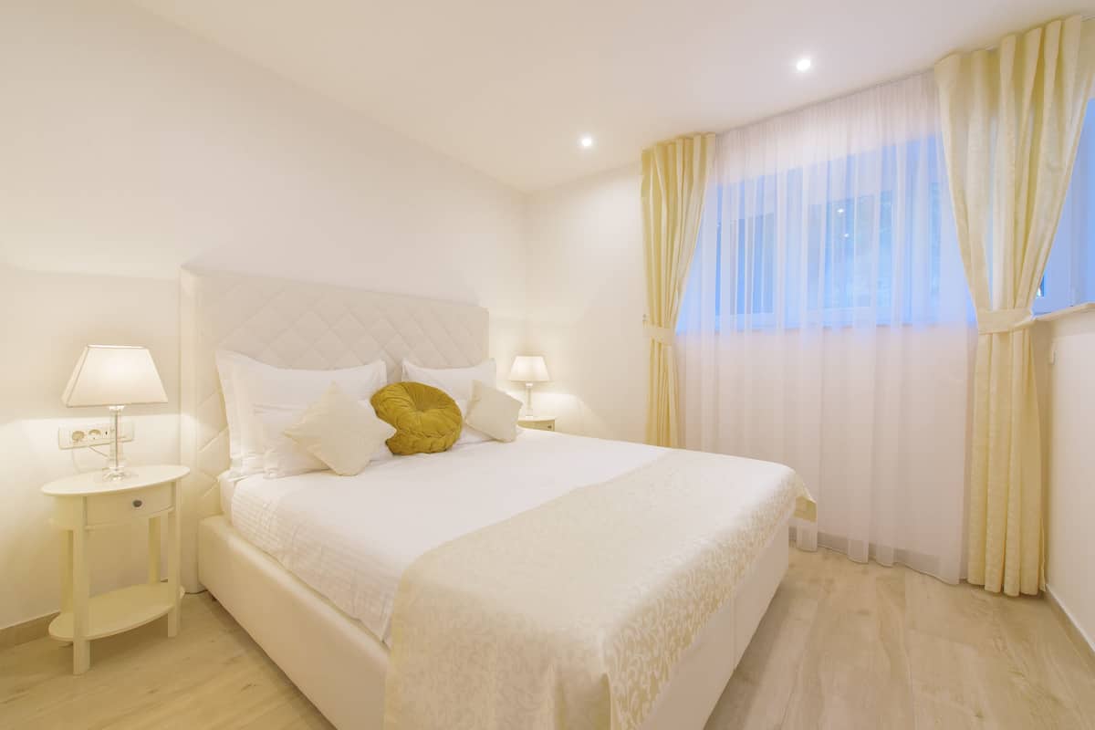 Quarto bem arrumado com uma cama grande, duas mesas de cabeceira com abajures e cortinas leves na janela.