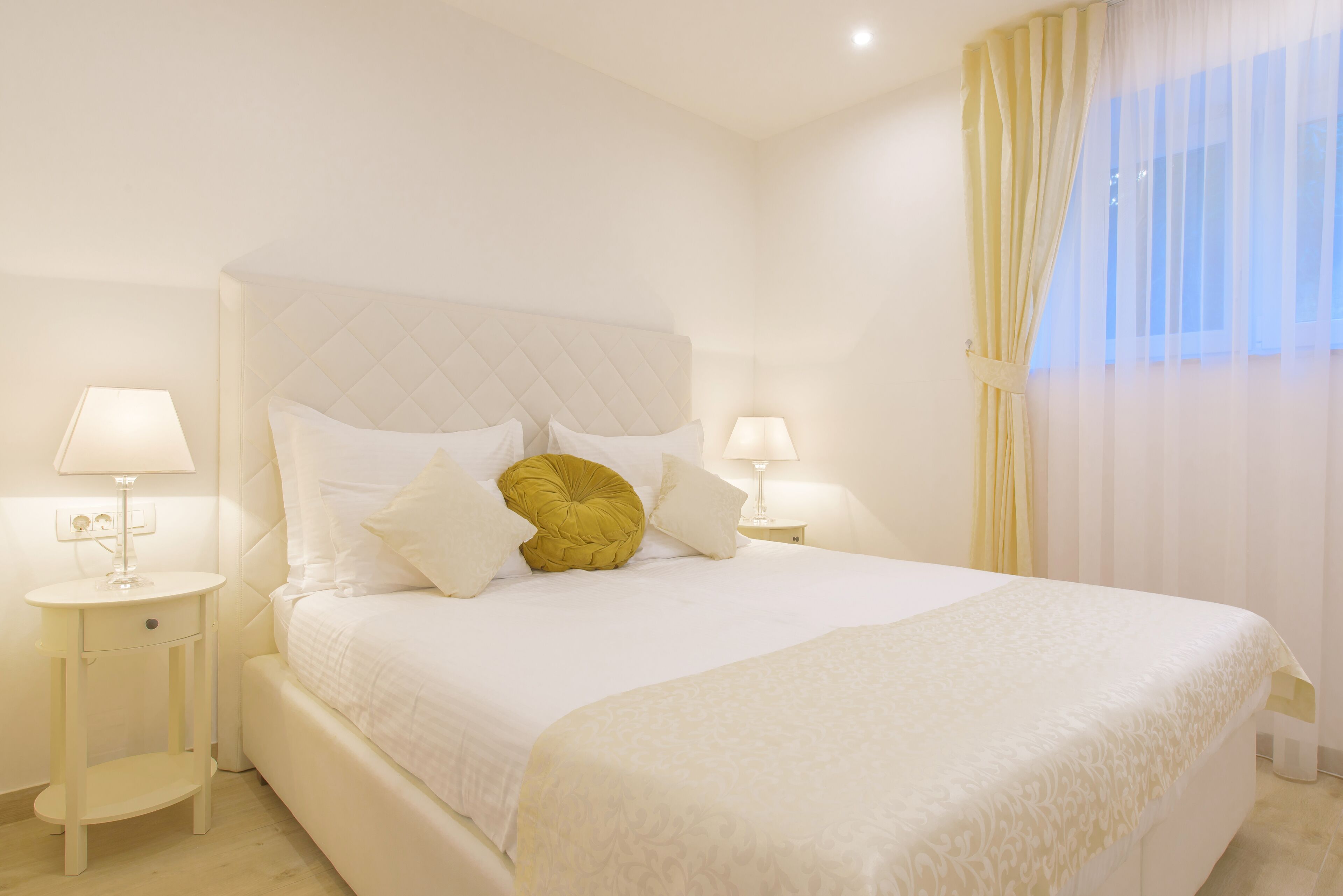 A neatly made bed with a quilted headboard, two bedside tables with lamps, and a window with sheer curtains.