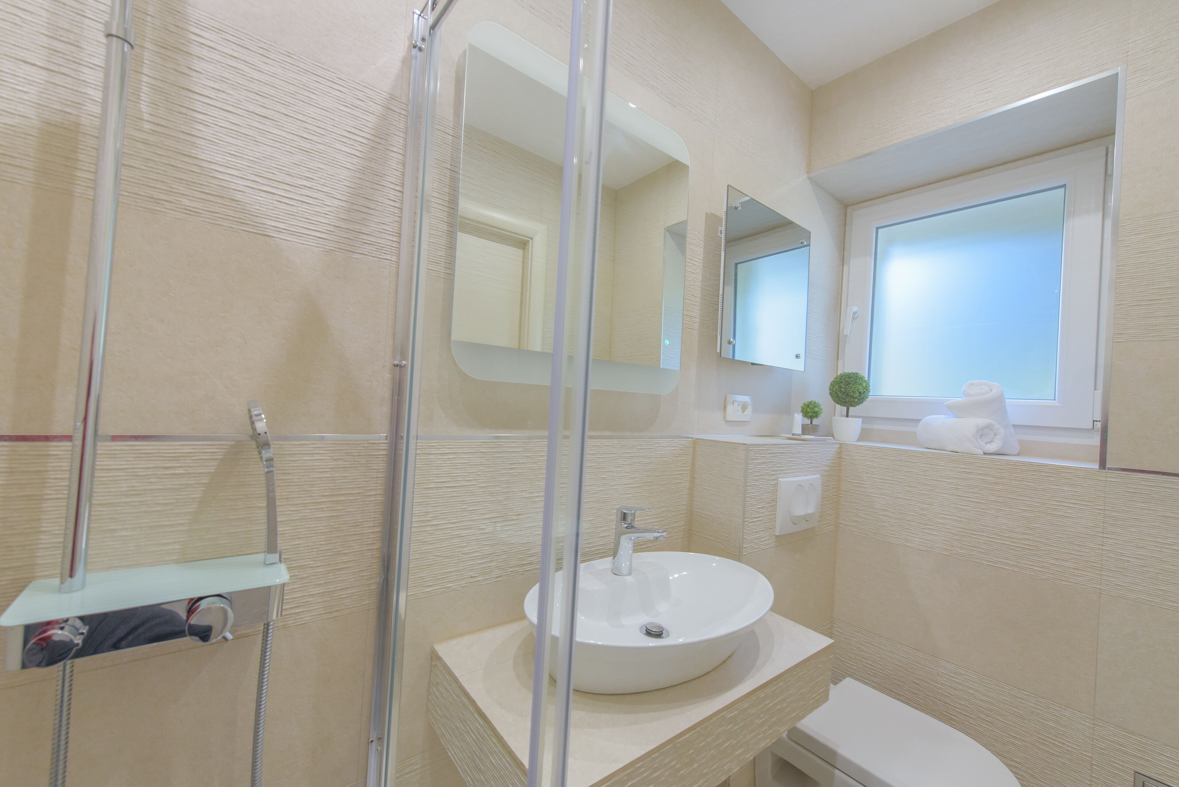 A modern bathroom with a glass shower enclosure, a sink with a mirror above it, and a window with a view.