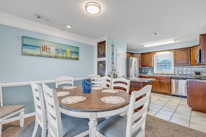 A dining area with a round table, white chairs, and a blue vase. There is a kitchen with wooden cabinets and a refrigerator.