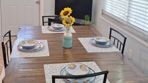 A dining room with a wooden table, chairs, and a vase with sunflowers. There is a television on the wall and a door with a window.